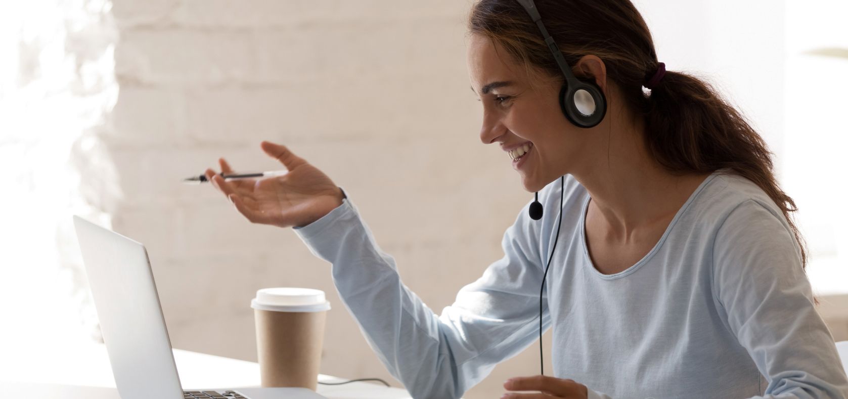 Smiling woman wearing a headset, talking and gesturing during a video call on a laptop with a coffee cup on the desk