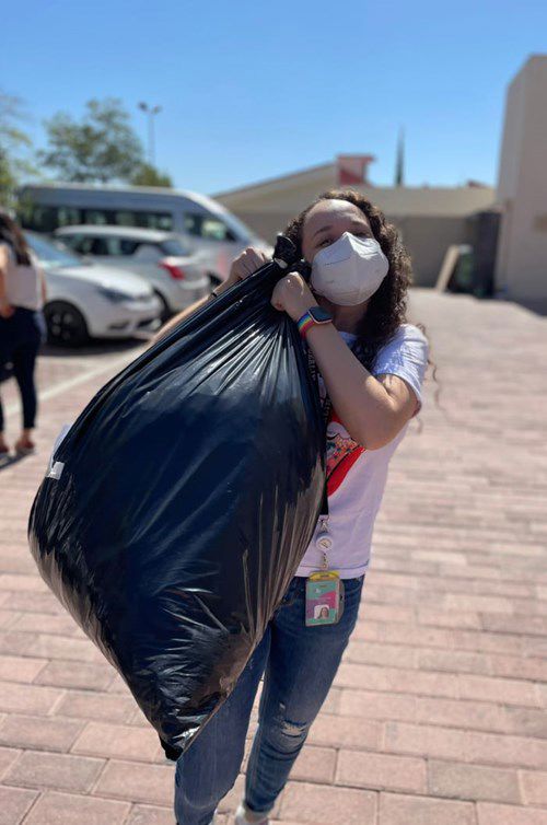 a person wearing a mask carries a large black garbage bag over their shoulder, walking on a paved area in a sunny outdoor setting with parked cars in the background. a person wearing a mask carries a large black garbage bag over their shoulder, walking on a paved area in a sunny outdoor setting with parked cars in the background.