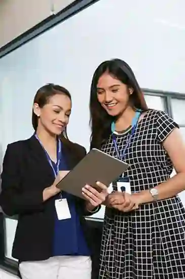 Two TP Asian female professionals working on a tablet; one wearing a long black checkered dress, the other in white pants and blue blouse.