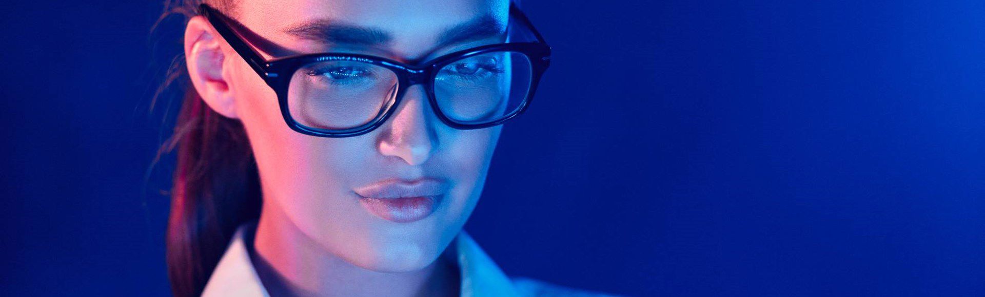A person wearing glasses and a white shirt, illuminated by blue and pink lighting, appears focused and engaged, set against a solid blue background. A person wearing glasses and a white shirt, illuminated by blue and pink lighting, appears focused and engaged, set against a solid blue background.