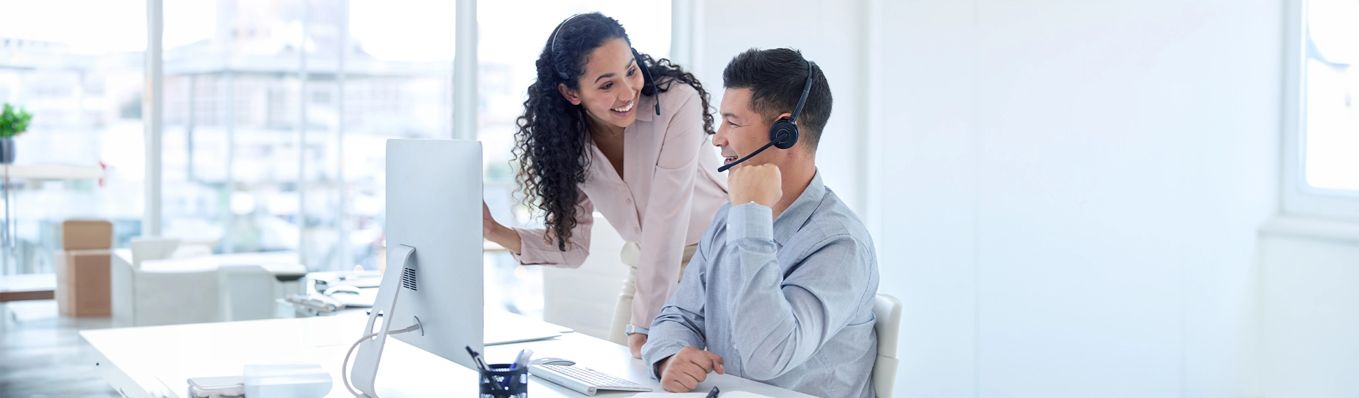 Two professionals collaborating in a modern office setting, discussing work at a desktop computer. One person is wearing a headset, and the workspace features bright natural lighting and minimalistic design.