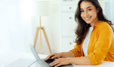 Person working on a laptop at a desk, wearing a yellow shirt in a bright home office setting.