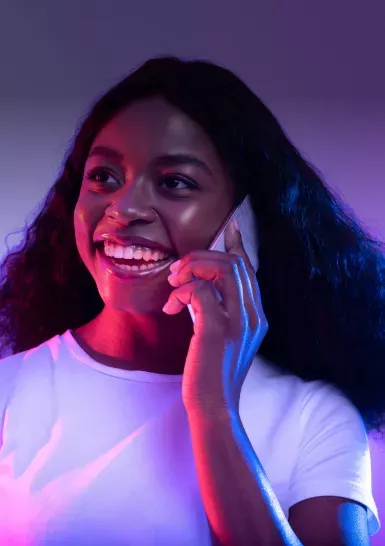 a woman smiles while talking on a mobile phone, wearing a white shirt. she is illuminated by vibrant pink and blue lighting in a simple studio setting.