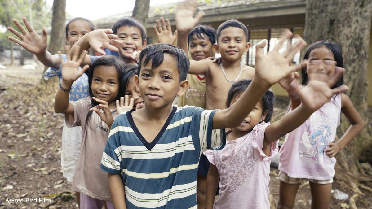 Children supported by the TP and UNICEF partnership smiling and waving at the camera.