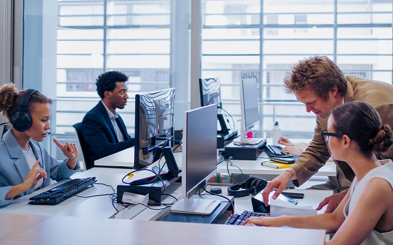 Modern office workspace with employees collaborating and working on desktop computers, featuring multiple monitors, headsets, and documents on a bright desk in a professional environment.