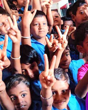 Children wearing colorful shirts raise their hands to make peace signs, smiling and gathered closely together, suggesting a cheerful group setting or event.