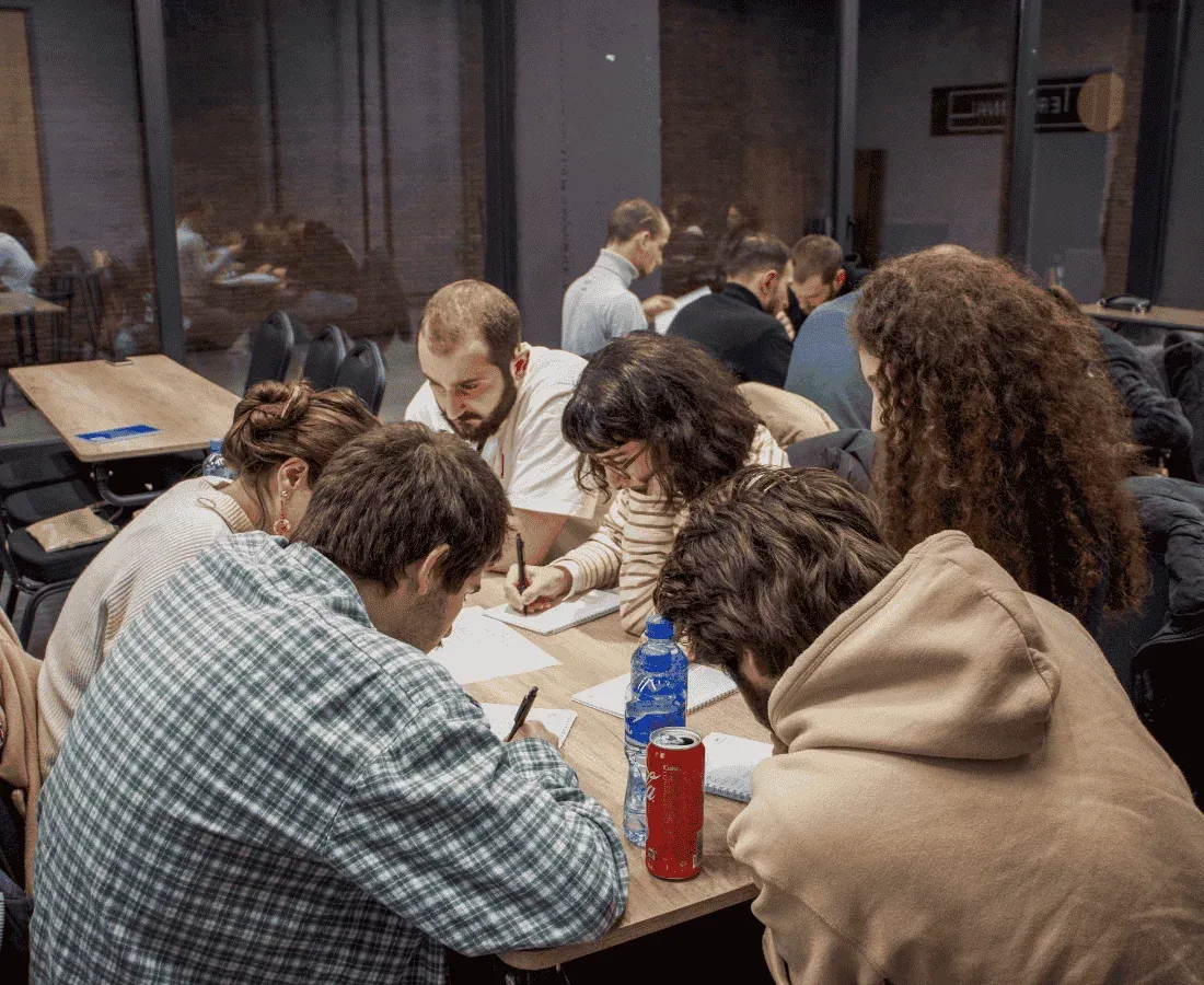 ჯგუფი ადამიანები სხედან მაგიდის ირგვლივ და ყურადღებით წერენ ქაღალდზე. მაგიდაზე დგას ბოთლები და ქილები. ისინი კლასში ან საკონფერენციო ოთახში იმყოფებიან.