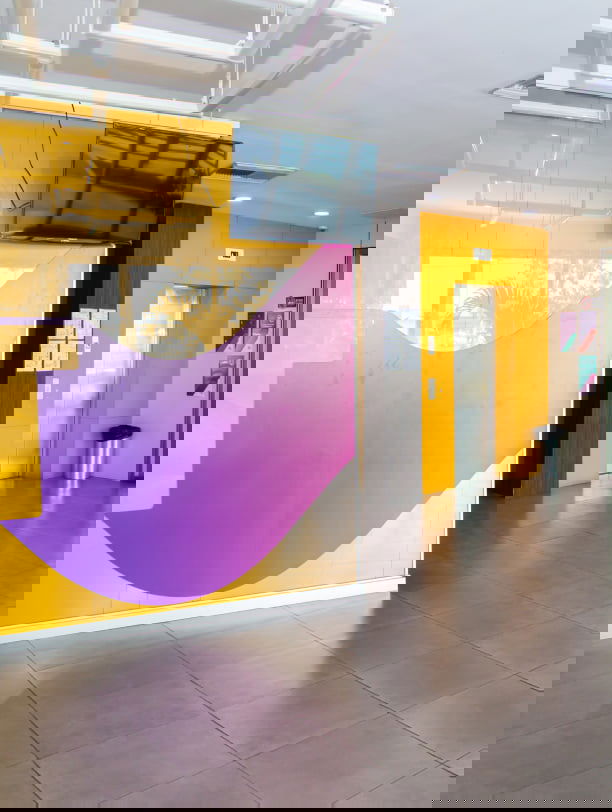 Área de recepción con paredes amarillas, un mural abstracto morado y pantallas montadas en la pared cerca de una puerta. Área de recepción con paredes amarillas, un mural abstracto morado y pantallas montadas en la pared cerca de una puerta.
