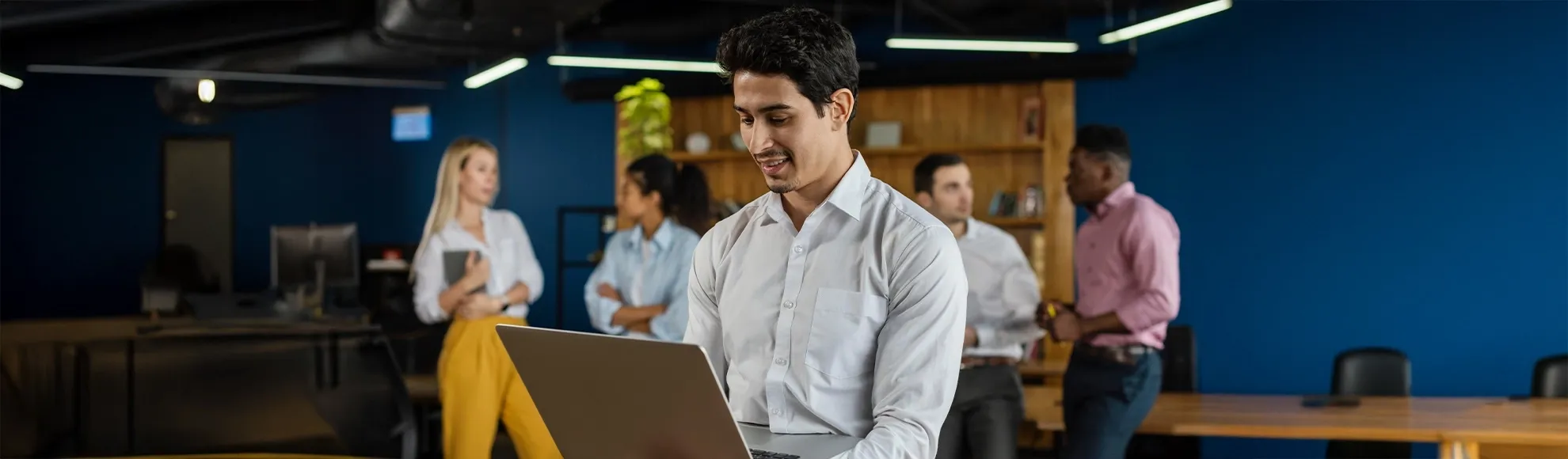 Professional man using a laptop in a modern office while colleagues talk in the background.