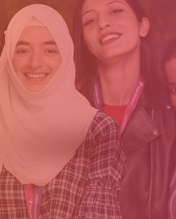 Two people smiling, one wearing a hijab, the other in a jacket, stand closely together in a warmly lit setting, suggesting a friendly or social environment.