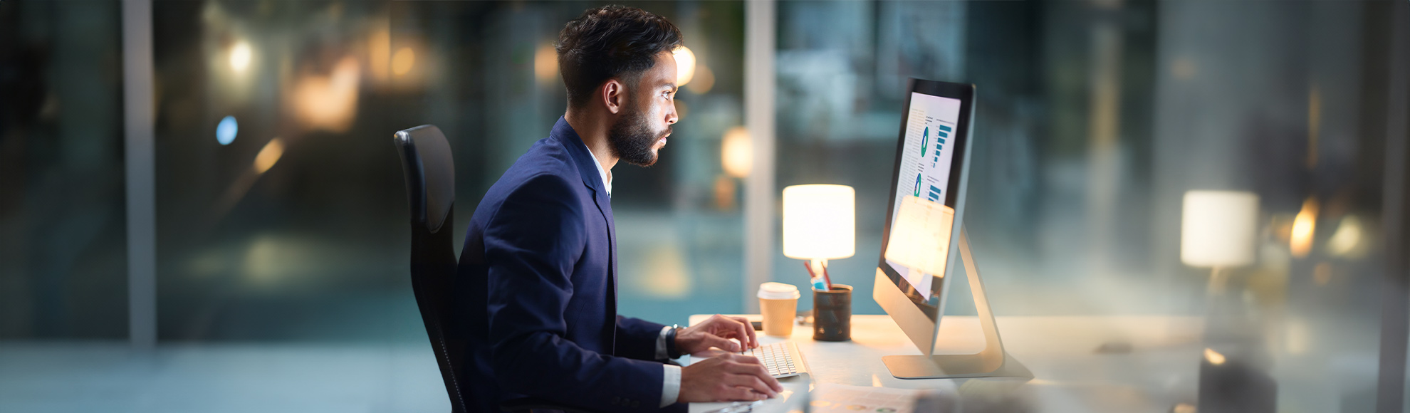 Business professional analyzing data charts on desktop computer in modern high-rise office with city view. 
