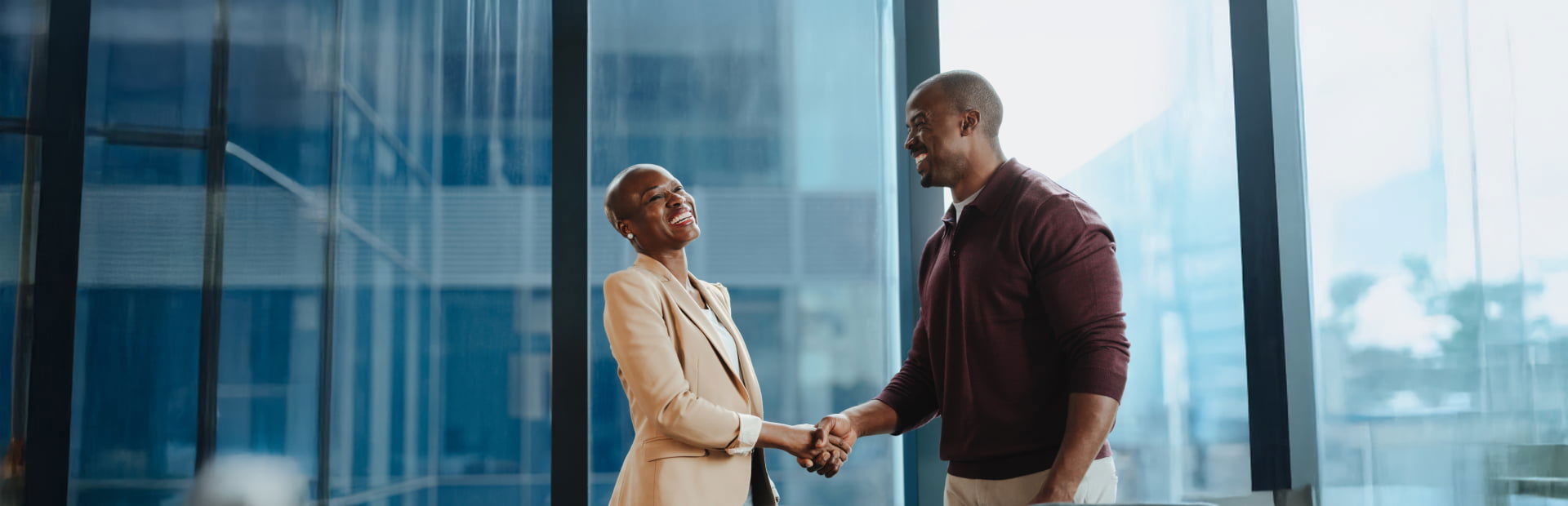 Two professionals smiling and shaking hands in a modern office with large glass windows