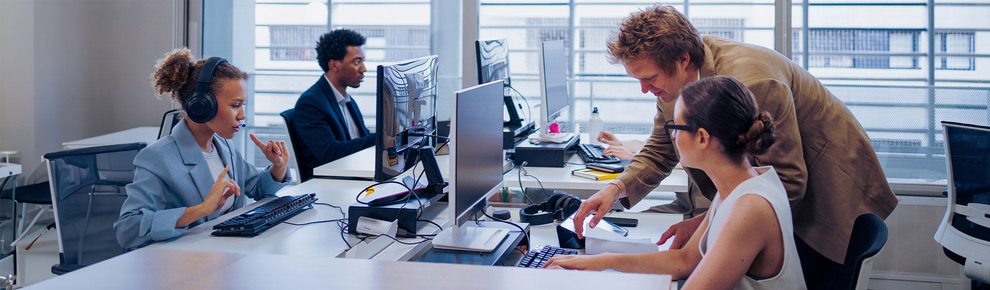 Modern office workspace with employees collaborating and working on desktop computers, featuring multiple monitors, headsets, and documents on a bright desk in a professional environment.