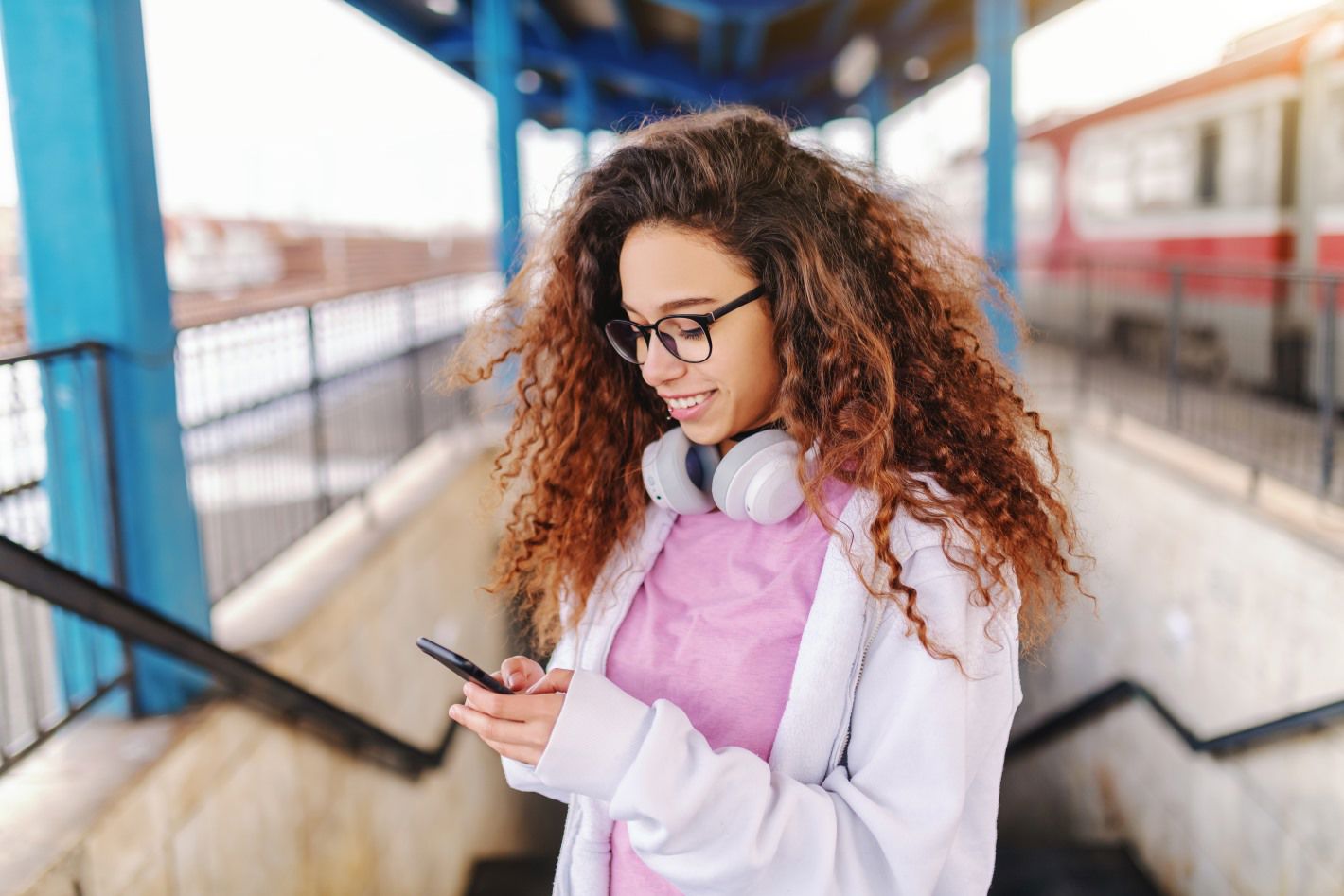 Young woman smiling while using her phone at a train station, wearing headphones around her neck.