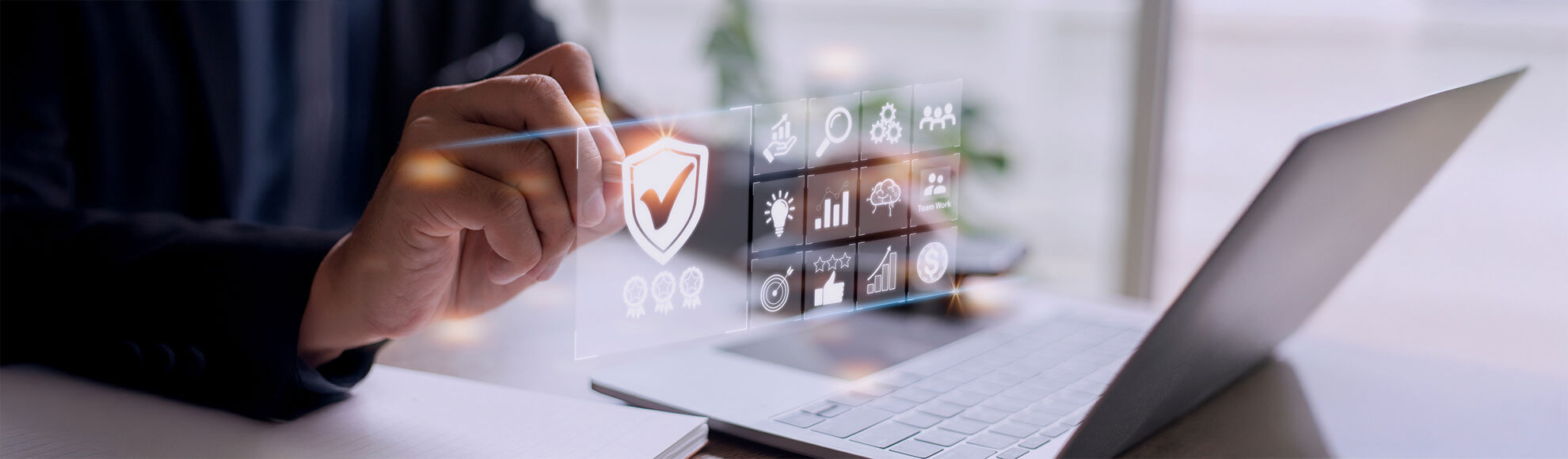 Person interacting with a floating digital interface above a laptop, featuring icons for cybersecurity, data analytics, cloud computing, and user management, including a shield with a checkmark symbolizing security.