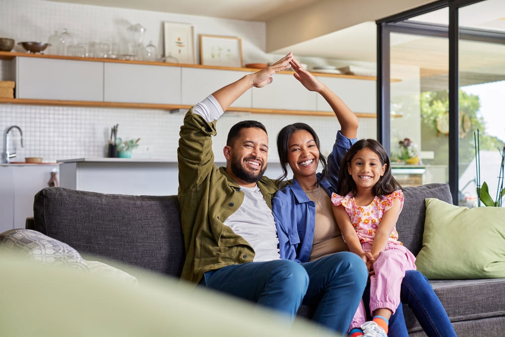 Smiling family sitting on a sofa at home, with parents forming a house gesture, representing TP’s Insurance page.
