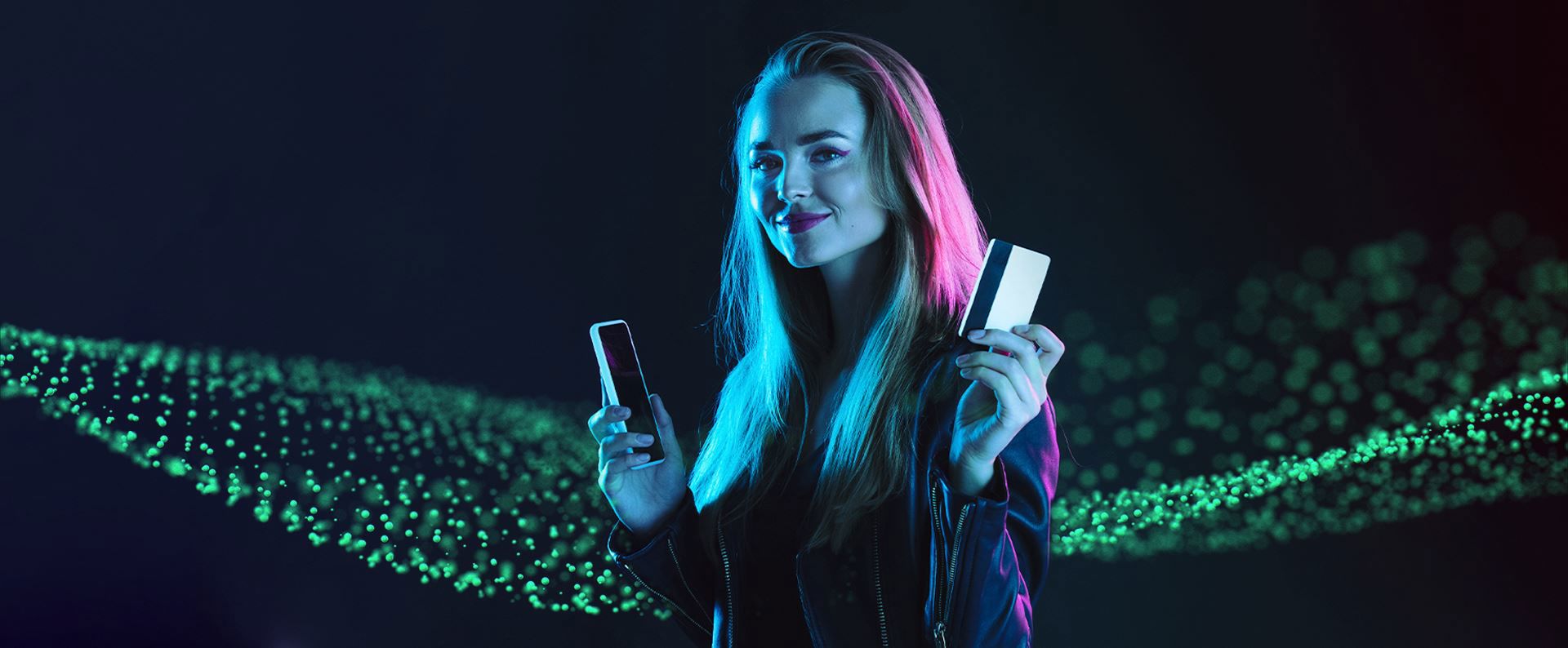 a person holds a smartphone and a credit card, smiling under blue and pink lighting against a dark background with scattered green light particles.