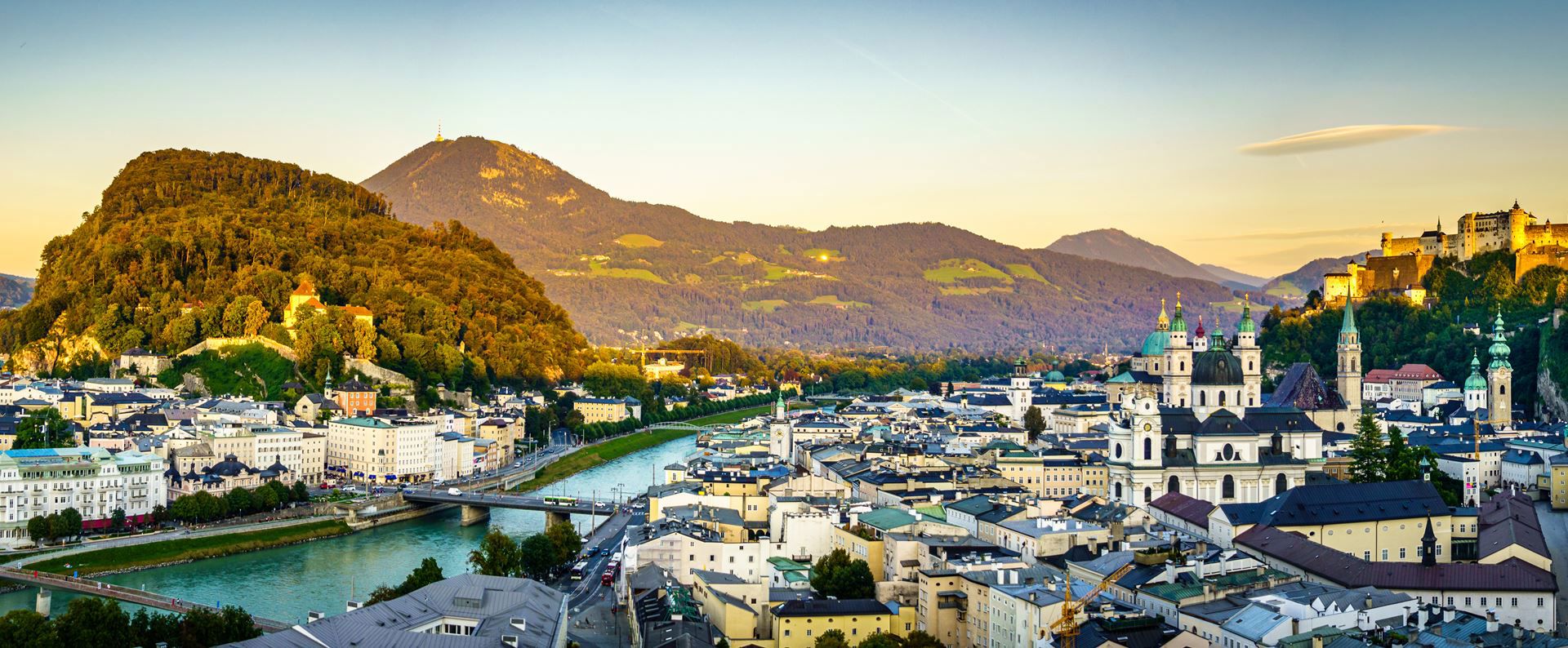 Cityscape of Salzburg with river, historic buildings, churches, and fortress against a mountainous sunset.