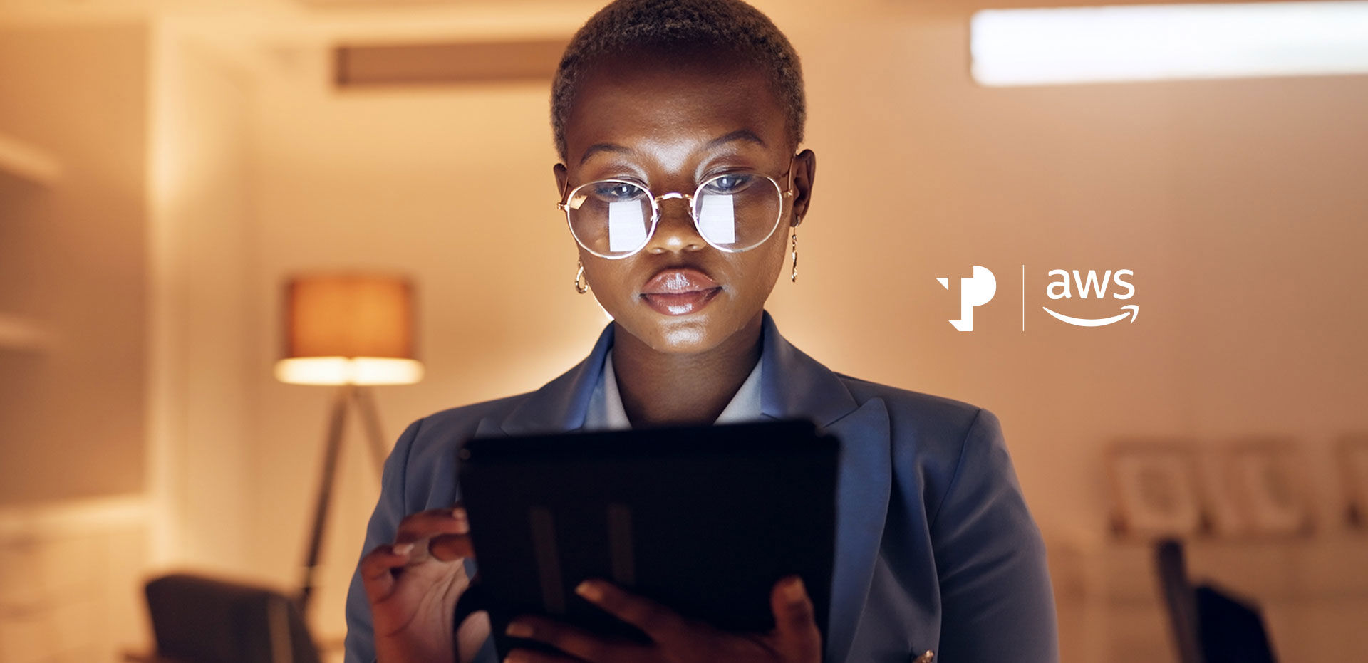 A person with glasses reads a tablet in a warmly lit room with a floor lamp. logos in the image: "p" and "aws."
