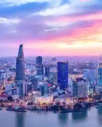Skyscrapers stand tall reflecting city lights along a river, under a vibrant sunset sky, creating a colorful urban skyline in a bustling metropolis.