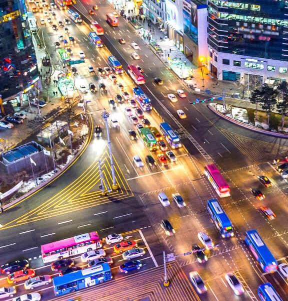 자동차와 버스들이 밤에 불빛으로 가득한 분주한 도심 교차로를 지나가고 있으며, 주변은 높은 건물과 밝은 조명으로 둘러싸여 있습니다.