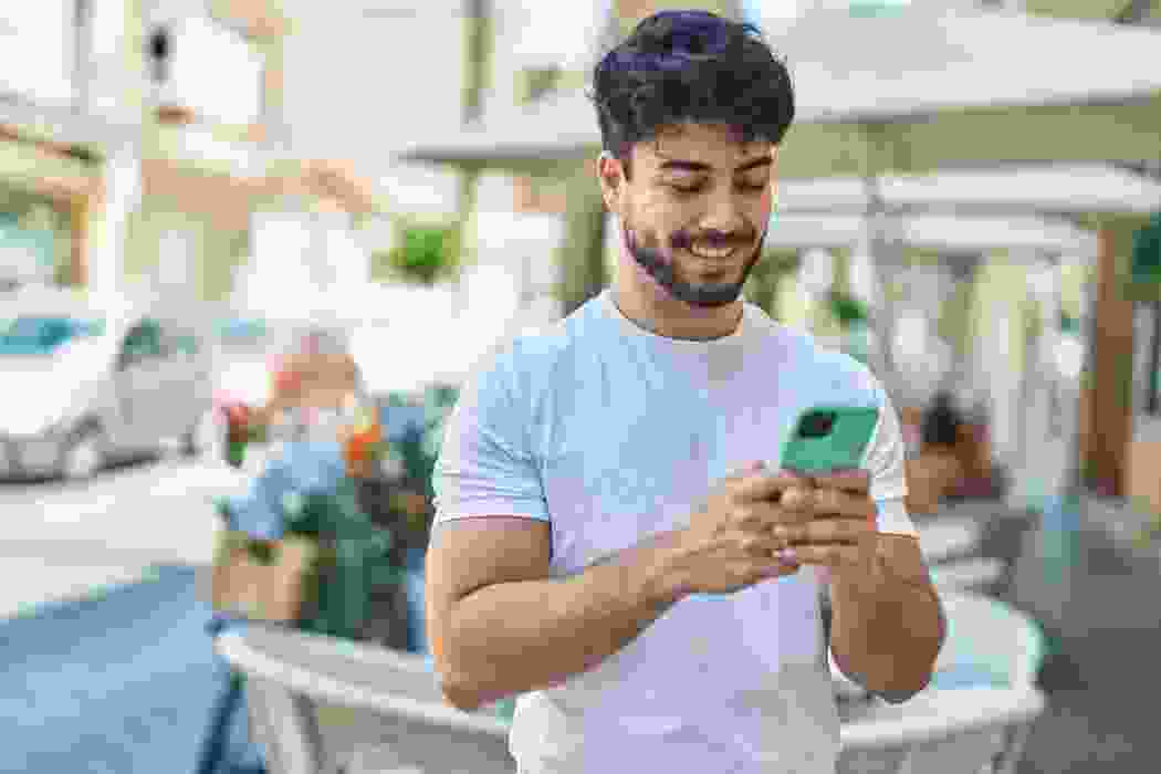 Young man in a white T-shirt holding his phone at an outdoor café on a sunny day.