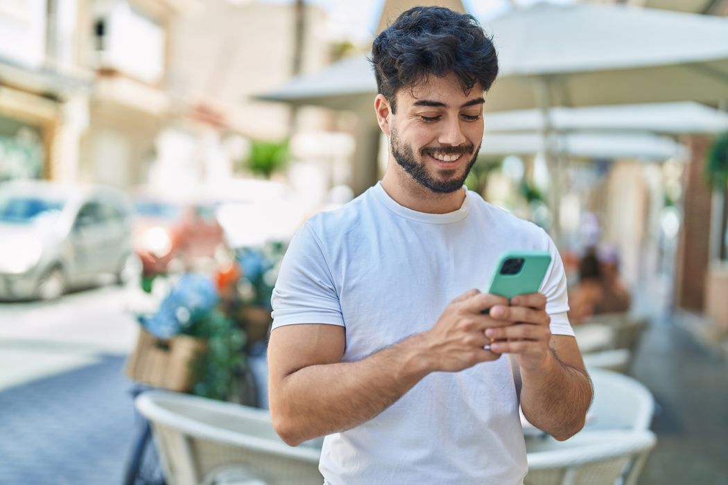 Young man in a white T-shirt holding his phone at an outdoor café on a sunny day.