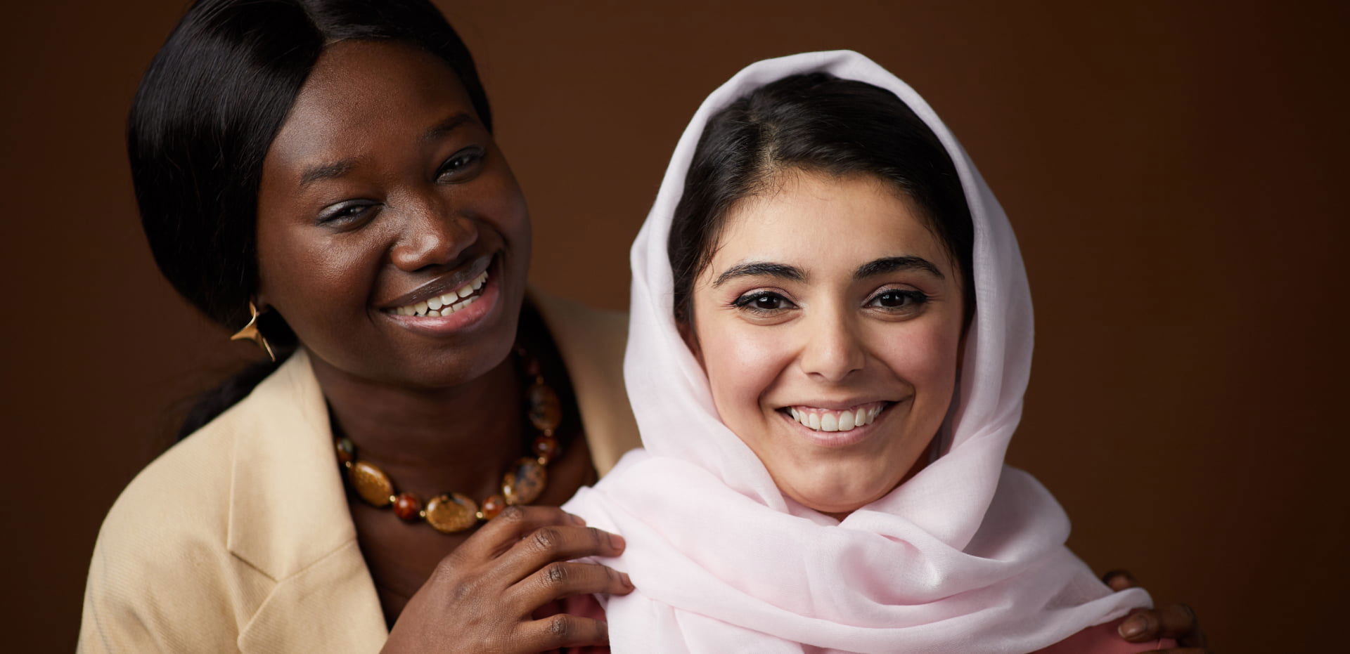 Two smiling women, one wearing a headscarf, posing together against a dark background.