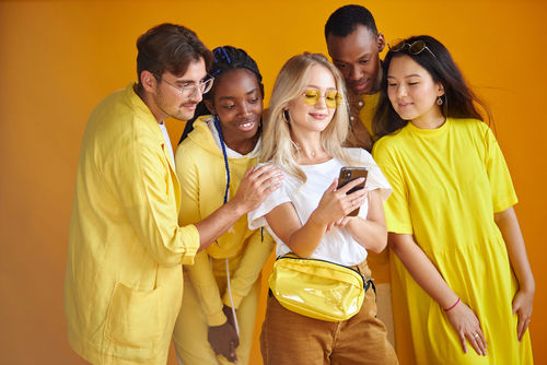 Group of diverse young people smiling and looking at a smartphone together against a yellow background Group of diverse young people smiling and looking at a smartphone together against a yellow background