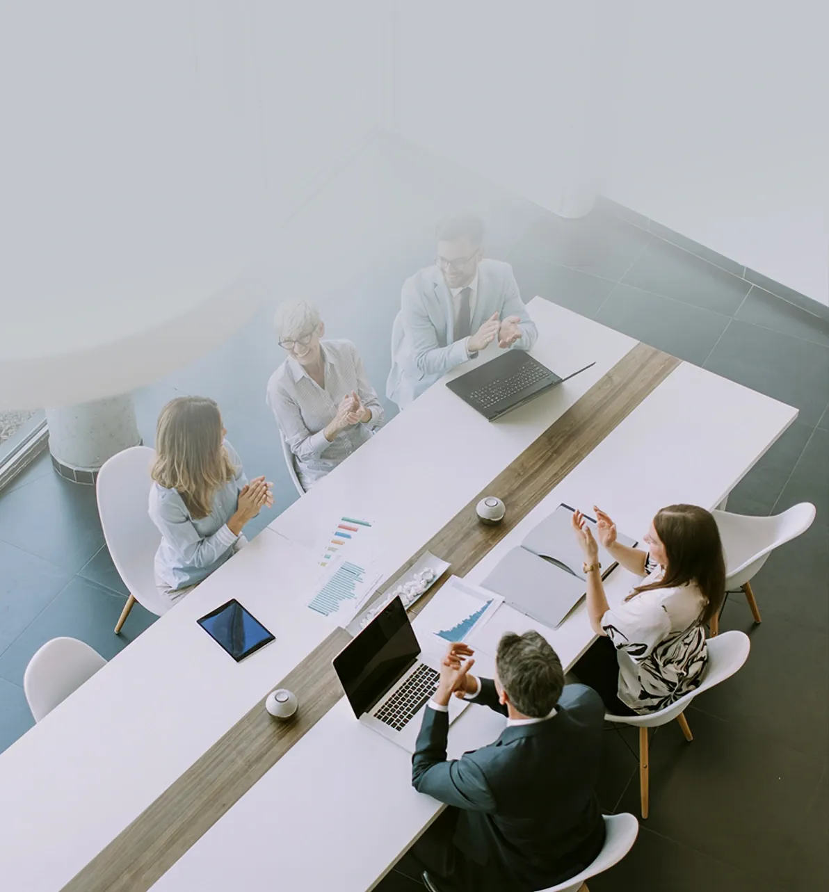 A team of business professionals claps together around a conference table, celebrating a successful meeting or milestone in a bright modern office. 