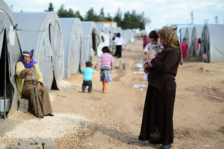 Row of tents in a refugee camp with people walking, sitting, and children playing.