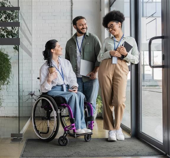 Three colleagues smiling in an office, including a woman using a wheelchair.