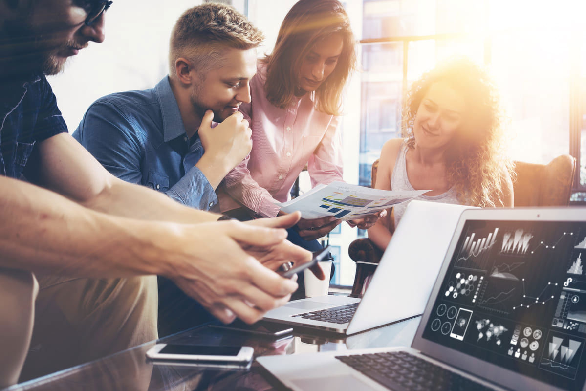 Four people sat around a desk with laptops, reviewing data performance and discussing the reports in the woman's hand