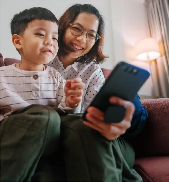 Mother and child smiling while using a smartphone together on a sofa.