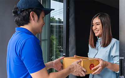 Courier in a blue uniform delivering a cardboard package to a customer at the doorstep of a modern home.