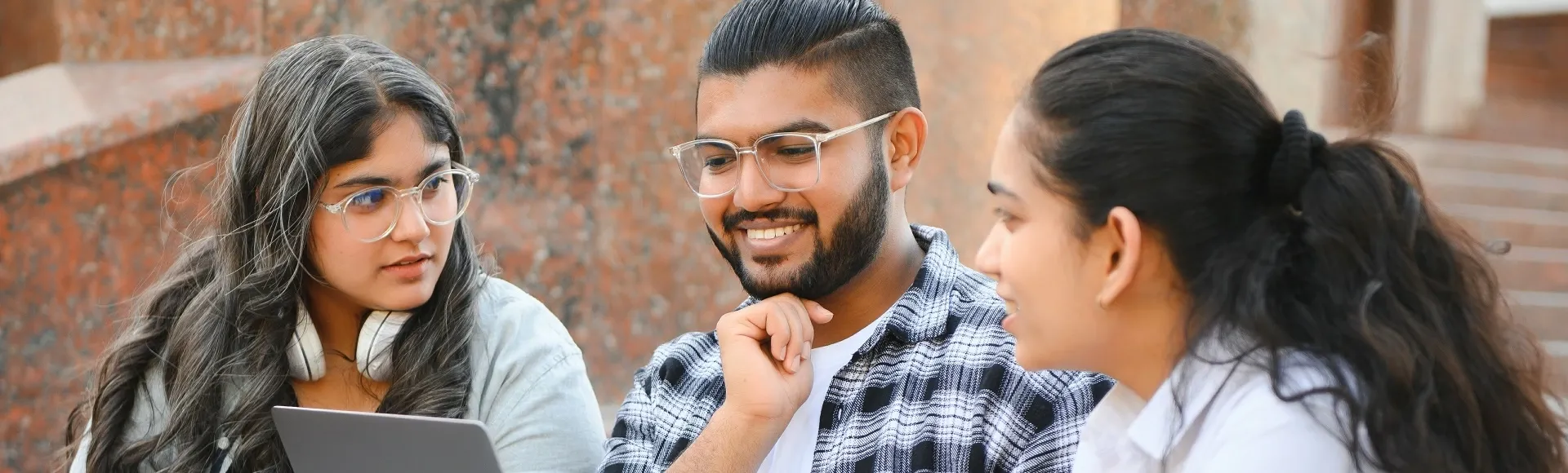 Three students collaborate outdoors, reviewing content on a laptop.