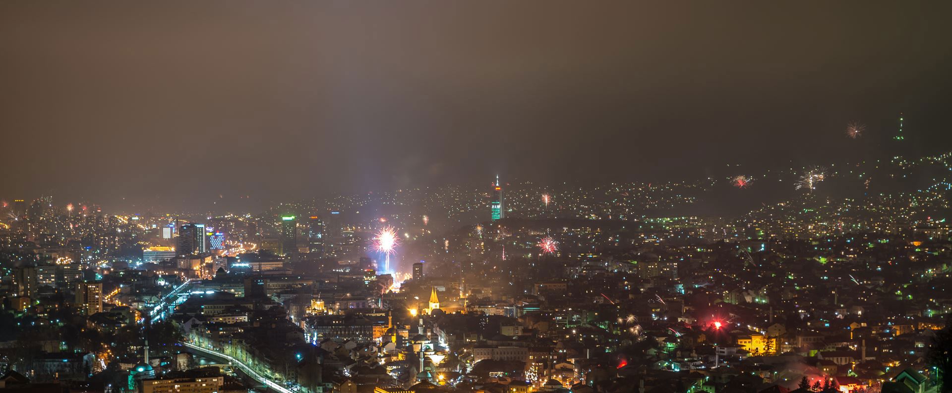 Vatromet iznad osvijetljenog gradskog pejzaža noću, s raznobojnim svjetlima koja obasjavaju visoke zgrade i ulice.
