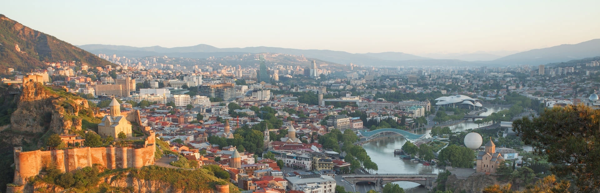 თბილისის ქალაქის ხედები გორაკზე მდგომი ციხესიმაგრით და ფერადი შენობებით, რომლებიც ველზეა გაშლილი.