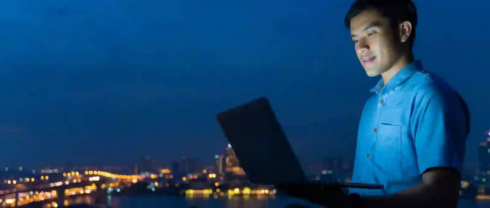 Man in a blue shirt using a laptop outdoors at night with a city skyline illuminated in the background