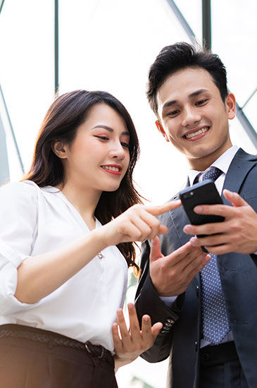 Two people are looking at a smartphone; one points at the screen while the other holds it, standing in a modern, bright indoor setting with geometric window patterns.