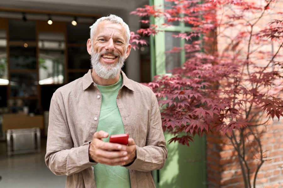 Smiling senior man holding his smartphone with flowers in the background, illustrating healthcare.