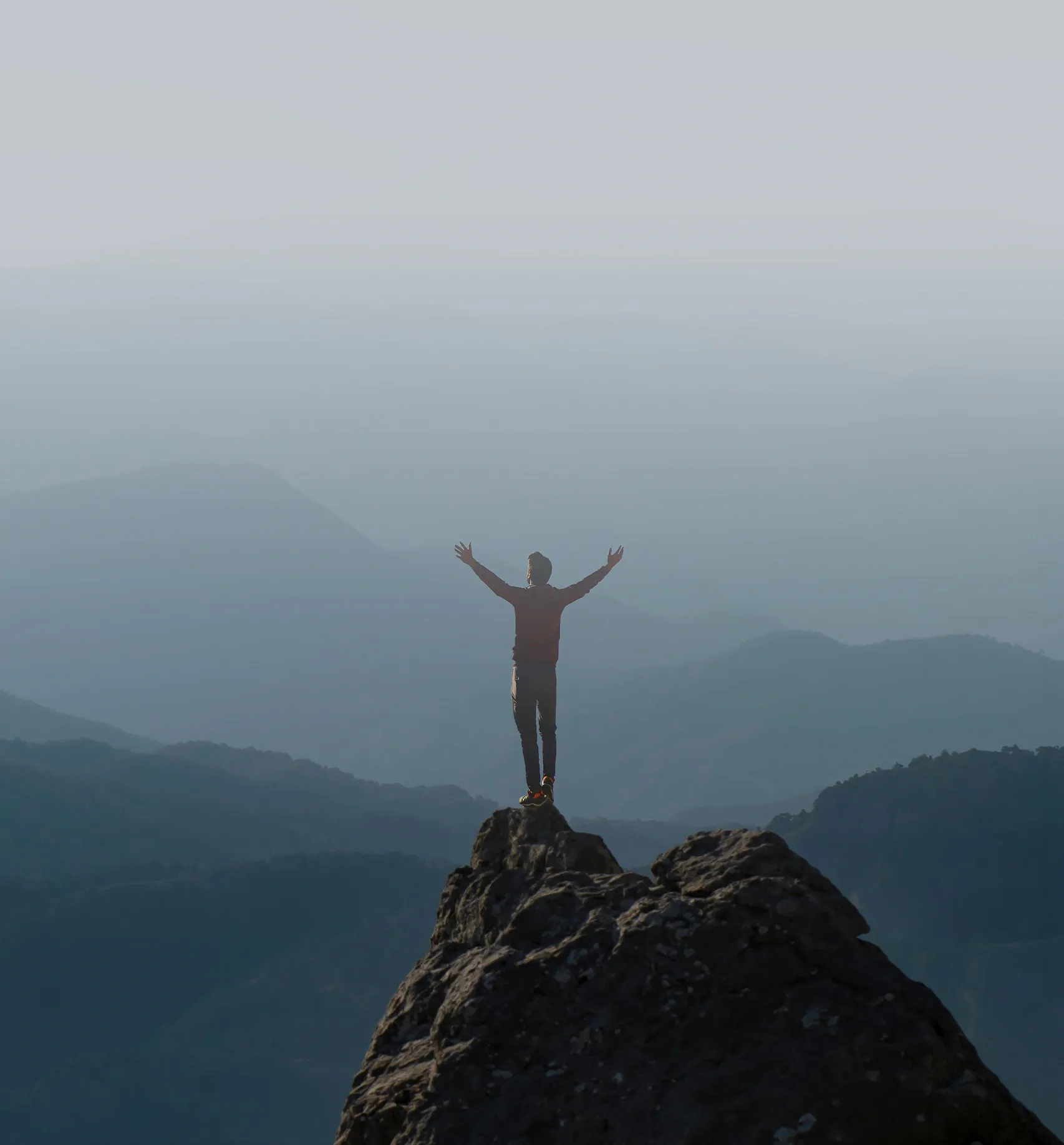 A lone hiker stands atop a rocky peak with arms outstretched, embracing the vast, mist-shrouded mountain ranges stretching into the distance. 