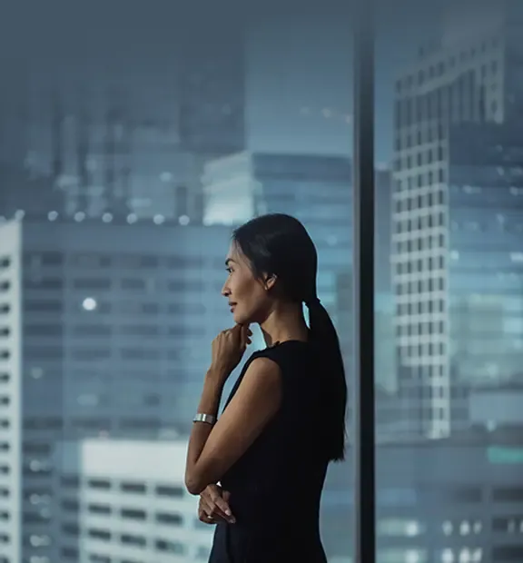 沉思中的亚洲女商务人士站在现代化办公室内，凝视窗外。