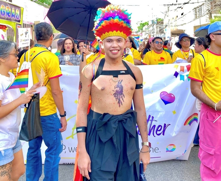 TP Pride parade participant in colorful headdress leading group with banner