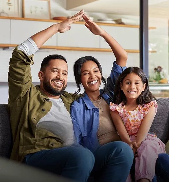 Smiling family sitting on a sofa at home, with parents forming a house gesture, representing TP’s Insurance page.
