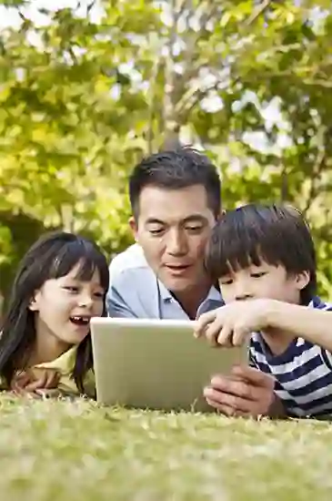 Asian family enjoying time outdoors in a park, reading stories together on a tablet.