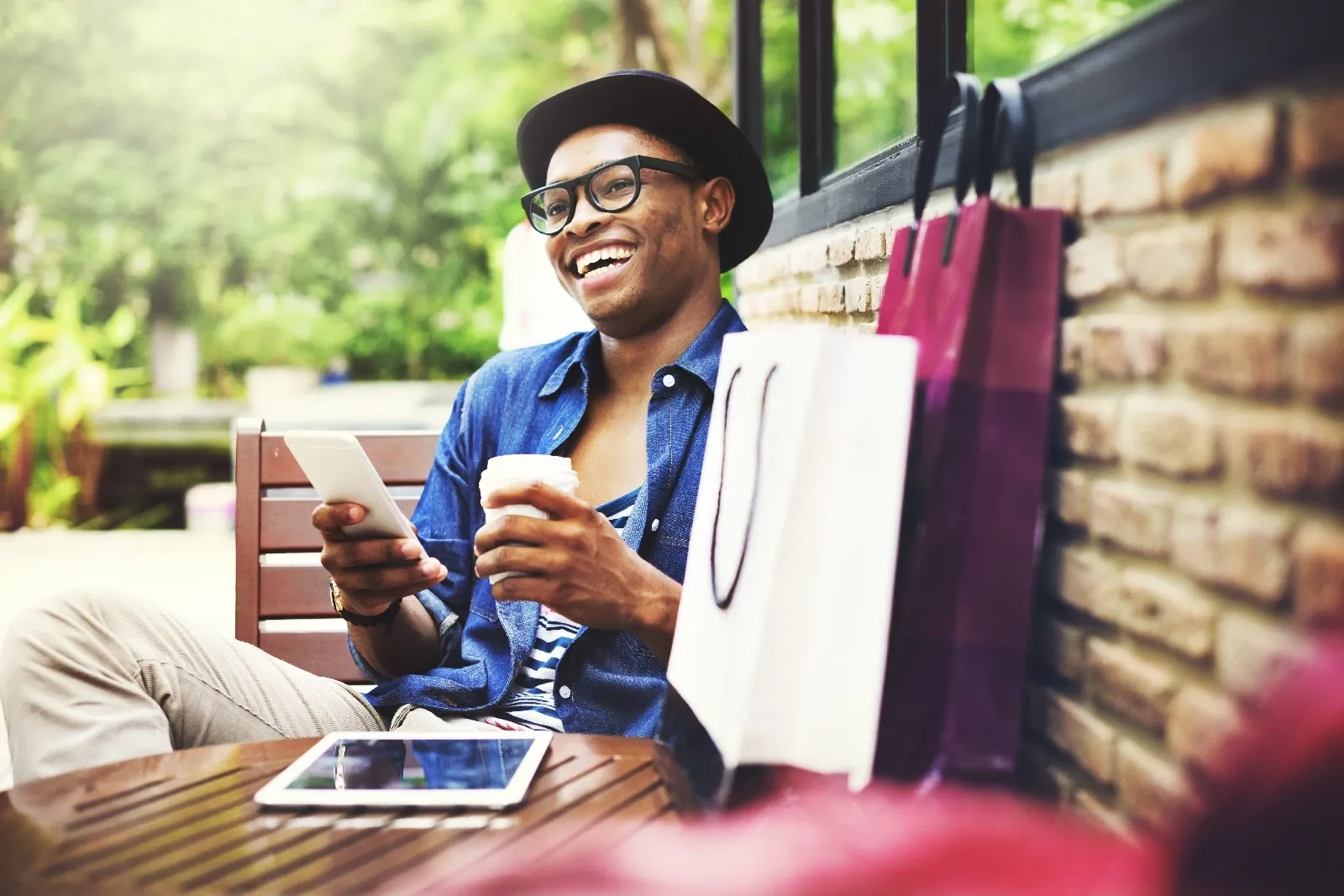Smiling man wearing hat and glasses holding coffee and tablet outdoors with shopping bags beside him.