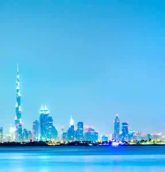 Dubai skyline with the Burj Khalifa and illuminated skyscrapers reflected on calm water under a clear blue sky.