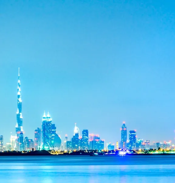 Dubai skyline with the Burj Khalifa and illuminated skyscrapers reflected on calm water under a clear blue sky.