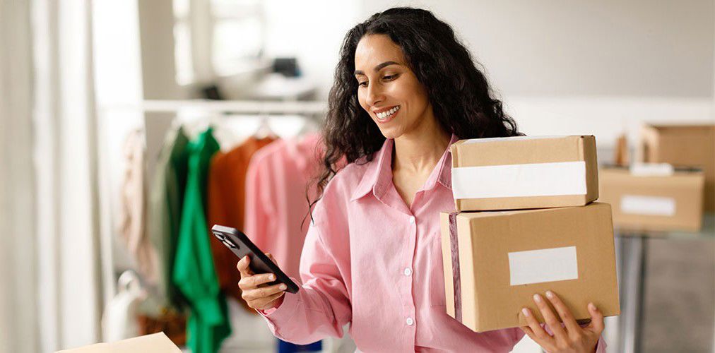A smiling woman in a pink shirt holding two cardboard boxes and using her smartphone, likely managing orders from home. The image represents efficiency and engagement in ad sales.